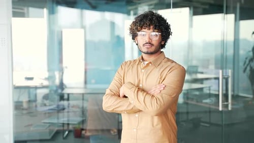 Portrait of young confident handsome man in glasses with crossed arms standing in modern office.