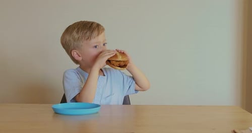 Blond Boy Enjoys Chicken Burger at Table Indoors