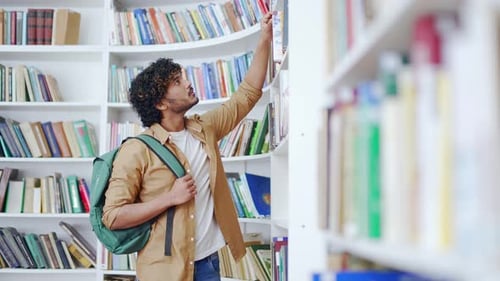 Student picking out a book in the library in the campus library space. A handsome male studying