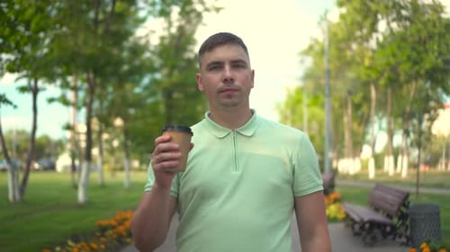 A Young Man Walks Along a Green Alley with Coffee in His Hands A Man Drinks Coffee and Walks in the