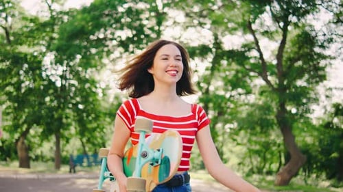 Beautiful Young Woman Walks Through the Park with a Skateboard in Her Hands The Girl is Carrying a