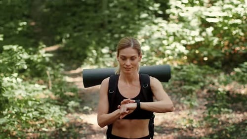 Woman Checking Watch While Hiking on Forest Path With Yoga Mat on Back