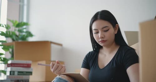 Woman Writing in Notebook Surrounded by Moving Boxes