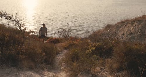 Hiker Man in a Tshirt and Shorts with Backpack Walking on the Coastline with Warm Sunset Light