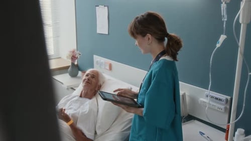 Young Medical Worker with Tablet Listening to Elderly Lady Lying in Hospital Bed