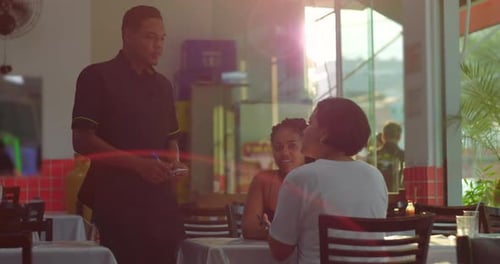Waiter writes down order at restaurant table while two women talk, sunlight flaring through large