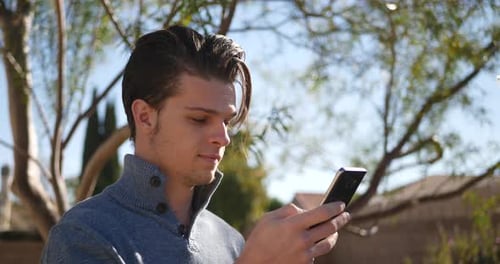 A handsome young millennial man in a sweater smiling and texting a friend on social media.