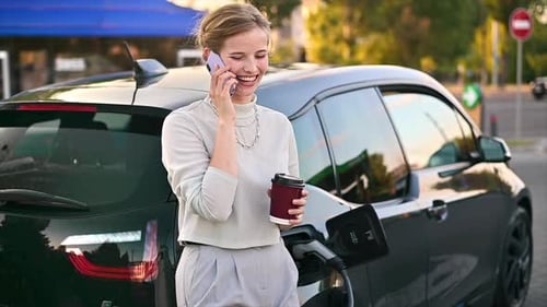 A young blonde woman with coffee talking on the smartphone at a car charging station with electric c
