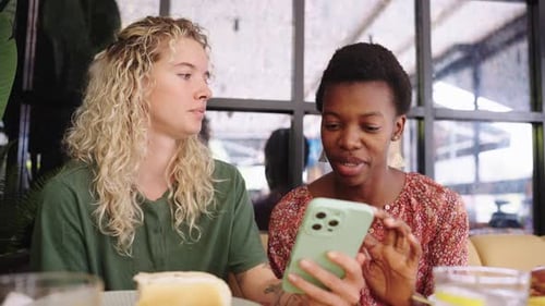Two Cheerful Diverse Women Using Smartphone and Laughing in a Cafe