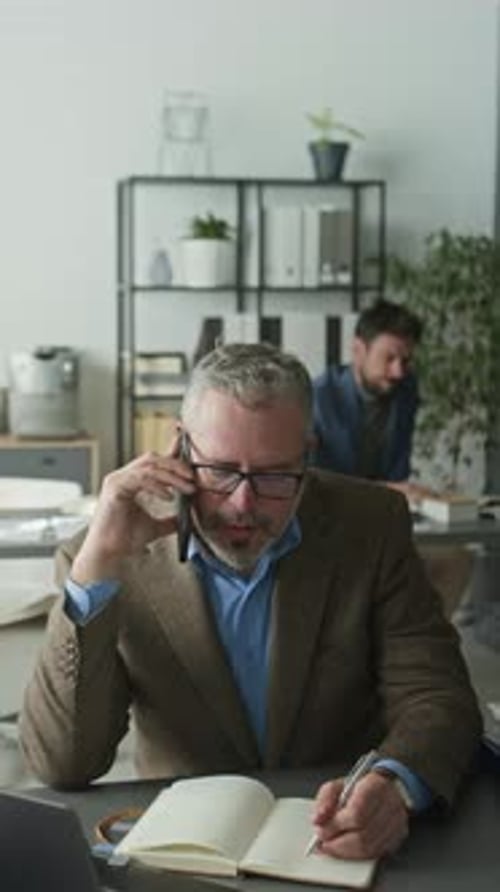 Businessman Having Phone Conversation and Taking Notes at Office Desk