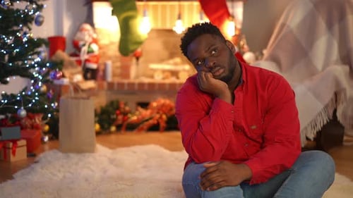 Sad Thoughtful African American Young Man Sitting in Living Room on Christmas Eve Looking Away