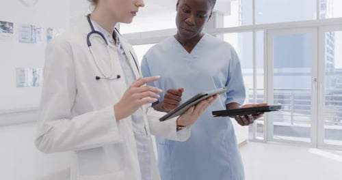 Diverse female doctor and nurse using tablets and discussing in hospital corridor, in slow motion