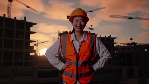 Asian Female Engineer Wearing Safety Helmet Smiling With Arms Akimbo At Building Site