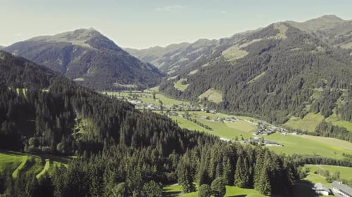 Aerial over picturesque tyrolean countryside with forest covered mountains and grassy fields. Houses