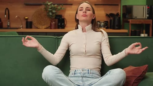 Young Brunette Meditates Sitting in a Lotus Position on a Green Sofa in the Living Room at Home A
