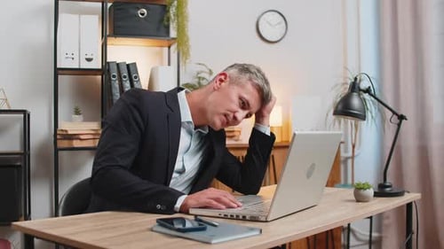 Exhausted Young Businessman in Formal Suit Working on Laptop Computer Sitting at Desk in Home Office