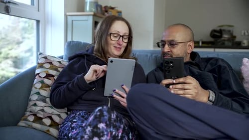 Interracial couple using tablet and smartphone sitting on sofa at home