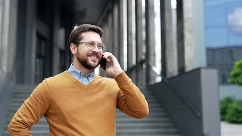 Smiling businessman talking on mobile phone while standing on street near office building. Handsome