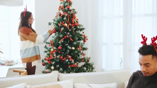 Multiracial Couple Celebrates Christmas As Woman Decorates a Festive Tree While Man Works on Laptop