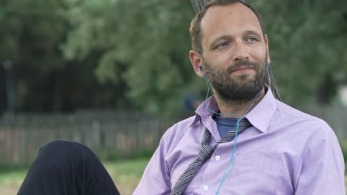 Young businessman enjoys music on his phone leaning against a tree in the park