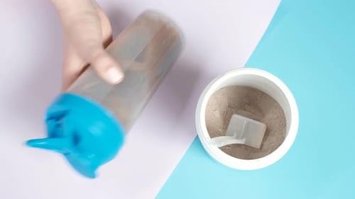 Female hand shaking bottle with a protein shake on a blue and pink background, top view.
