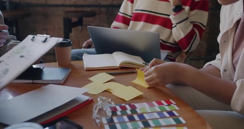 Nahaufnahme von Händen, die mit dem Laptop schreiben und Dokumente in der Hand halten, während Kollegen am Schreibtisch arbeiten