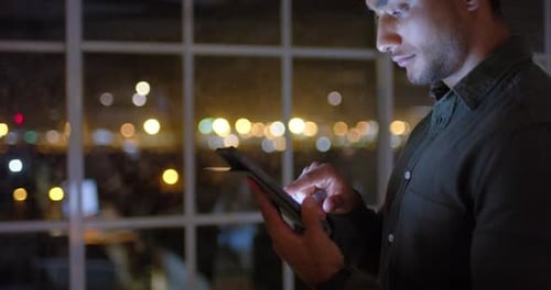 Thoughtful biracial businessman using tablet and looking out of office window at night, slow motion