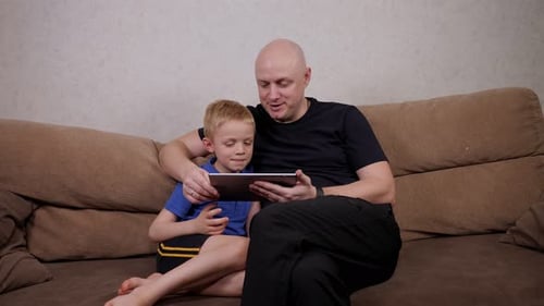 A Father and Son are Playing with a Digital Tablet Sitting at Home on the Sofa