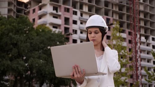 A Female Engineer in a Helmet with a Laptop is Standing at a Construction Site