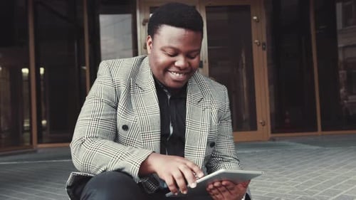 Cheerful AfroAmerican Man in a Formal Outfit Sits on the Stairs By the Office Entrance and Uses