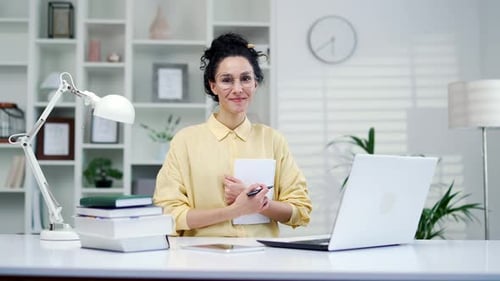 Portrait of young smiling female student with notebook in hands sitting at desk in home office. Posi