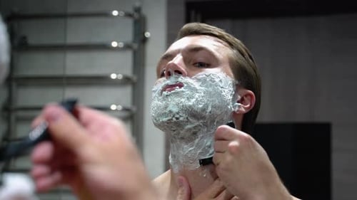 Man Shaves Beard with Razor in Bathroom Mirror