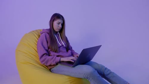 Young Woman Sitting on Pouf and Using Notebook Isolated on Purple Color Background Girl in Hoodie