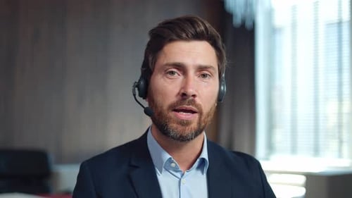 Bärtiger Mann mit Headset spricht während der Arbeit im Kundenservice-Center in die Kamera