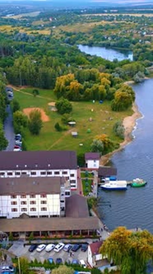 Green wooded bank of the river locating in the city. Hotel with the pier and few boats on the water.