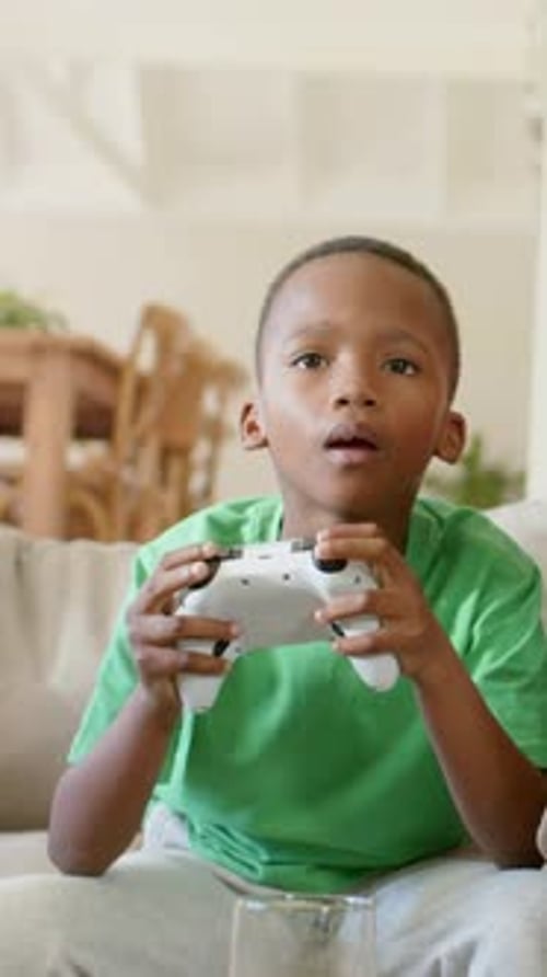 Vertikales Video: Ein Junge hält einen weißen Controller in der Hand und drückt Knöpfe beim Spielen auf der Couch in grünem Hemdglas