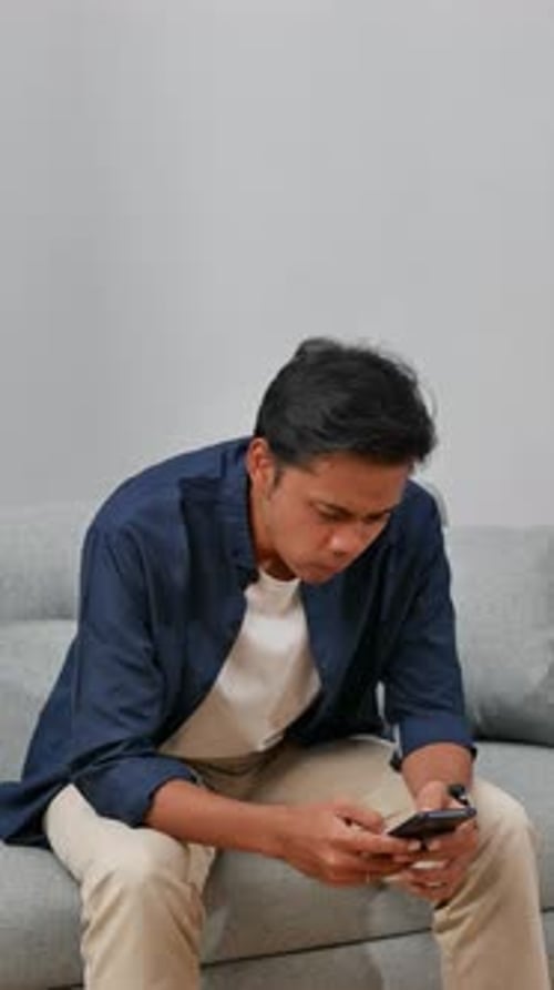 Pensive Upset Young Asian man Looking At Smartphone Screen At Home