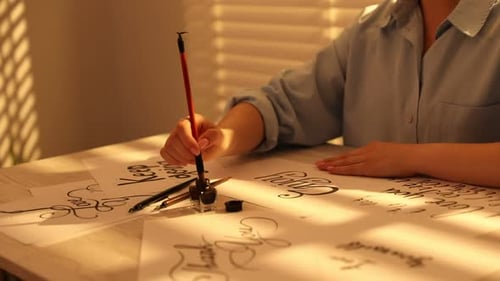 Kalligrafie. Frau mit Pinsel schreibt Wort Enjoy auf einem Blatt Papier am hellen Holztisch im Haus