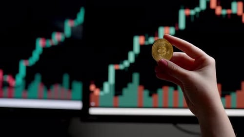 Halten Sie eine Kryptowährungsmünze in der Hand und beobachten Sie Marktdiagramme auf Computerbildschirmen