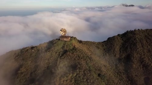 Aerial retreats from abandoned radar tower on mountain above clouds