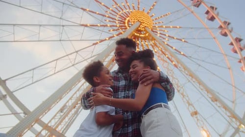 Optimistic Couple Hugging Son on Background of Ferris Wheel