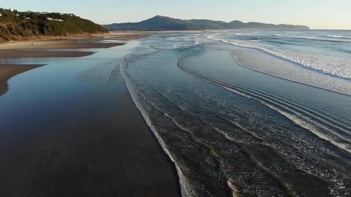 Drone flies over waves on the beach towards a mountain in the distance.