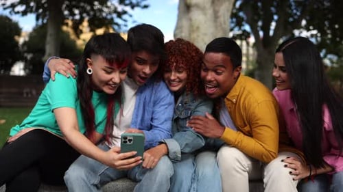 Multicultural group of friends sharing laughter while sitting on park bench, engaging with smartphon
