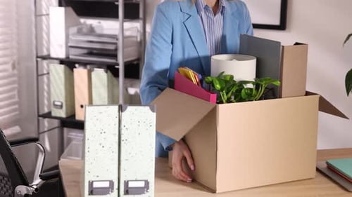 Dismissal. Woman packing her personal belongings into cardboard box at wooden table in office