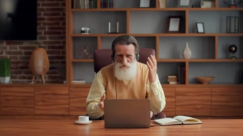 Older Man With Gray Beard Working on Laptop During Video Call at Home Office
