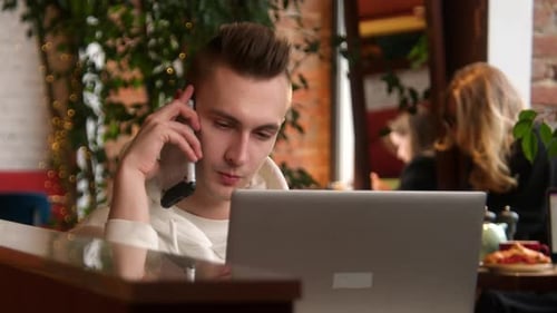Young Man is Talking on Phone at Laptop in Cafe Stock Footage Young Man is Talking on Phone While