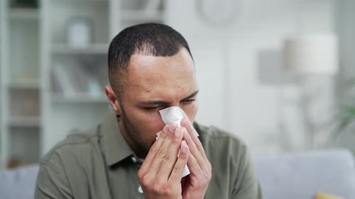 Ill young adult man sneezing in handkerchief blowing wiping running nose sneezing while sitting