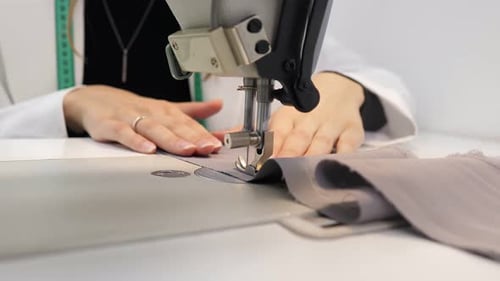 Woman Sews Grey Fabric on a Professional Sewing Machine While Sitting at Her Working Place in