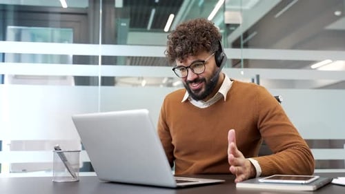 Confident businessman in a headset talking on a video call using laptop computer sitting in office.