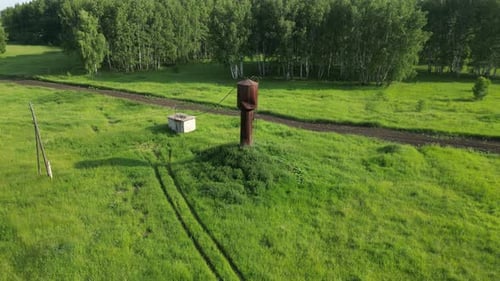 Weathered Water Tower Rising Above Verdant Meadow Bordering Dusty Road and Forest Edge Embodying